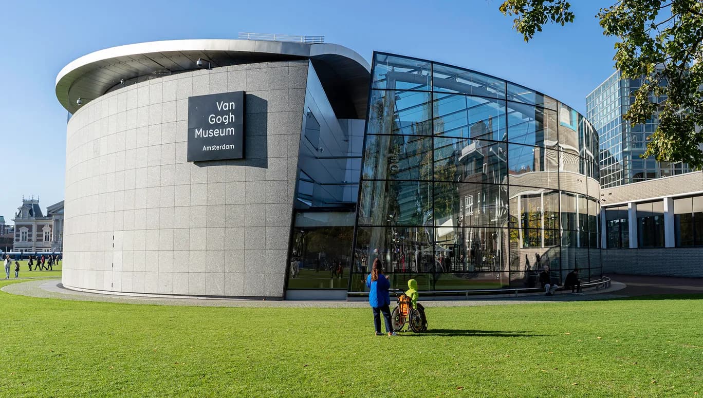 A person with a person in a wheelchair admiring the Van Gogh Museum at a sunny Museumplein.