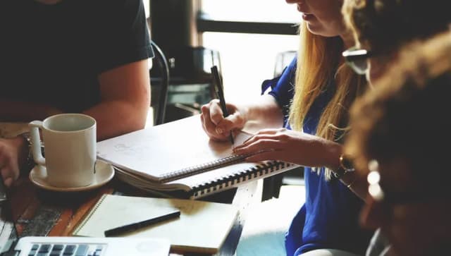 Closeup of hands taking notes on pad of paper next to coffee cup at desk during meeting.