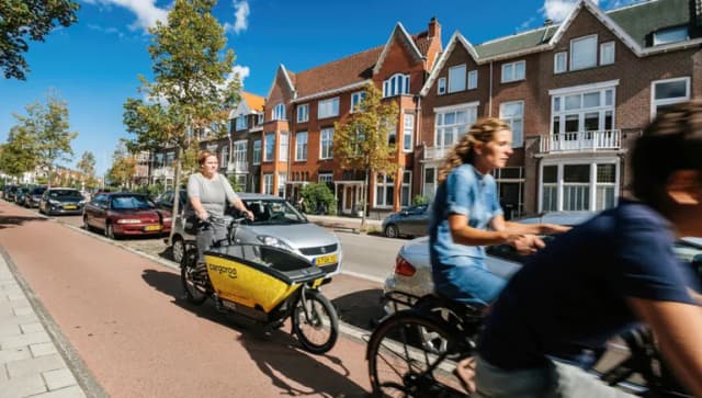 People riding bikes on Haarlem street with woman riding yellow shared Cargoroo e-cargo bike elektrische buurtbakfiets - fast commuting on Dutch street