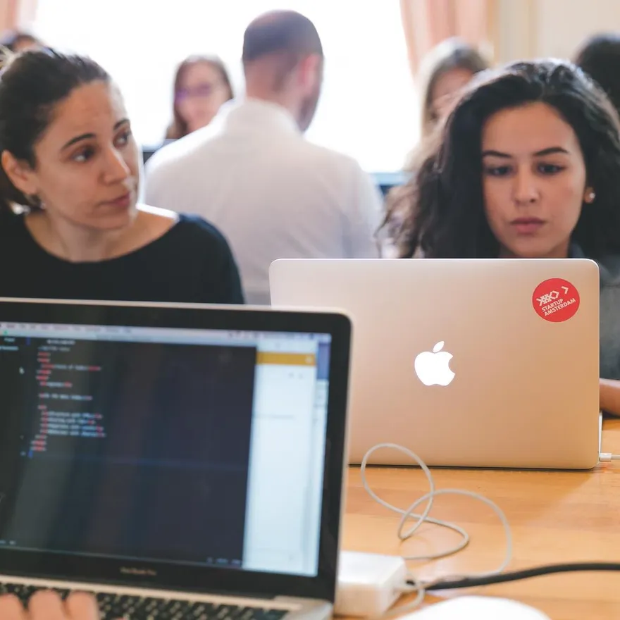 Closeup of students seated with laptops at table sat opposite each other.