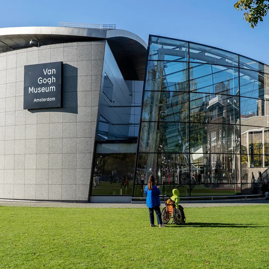A person with a person in a wheelchair admiring the Van Gogh Museum at a sunny Museumplein.