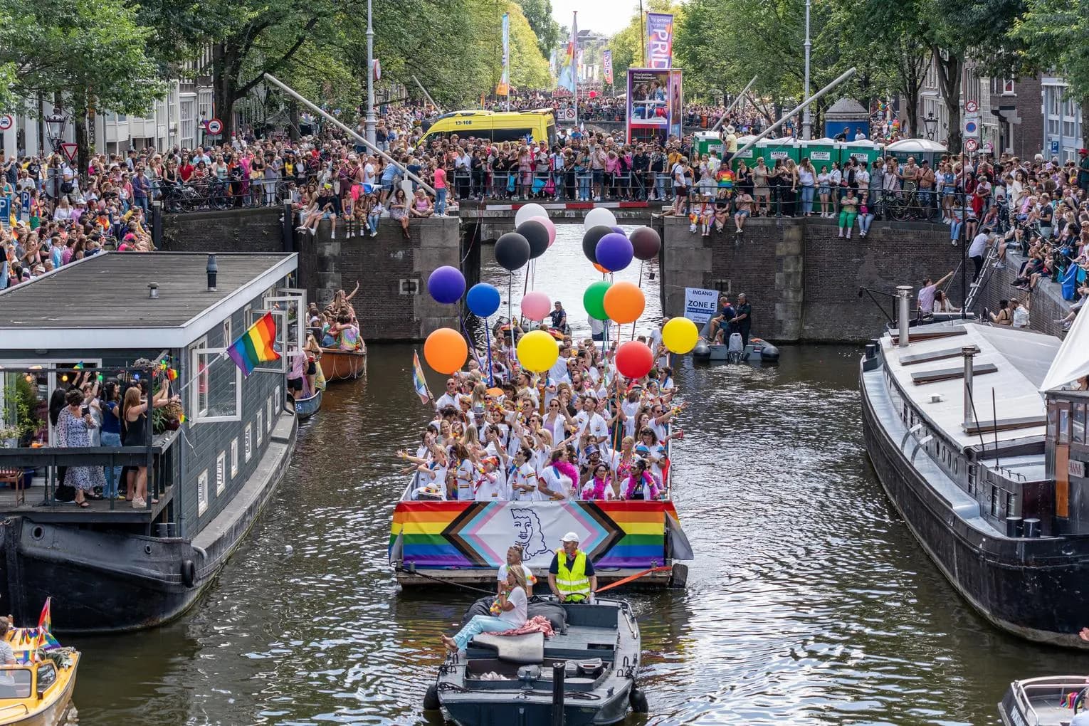 A crowd of people on the streets and in a boat with flags and signs - Pride Canal Parade